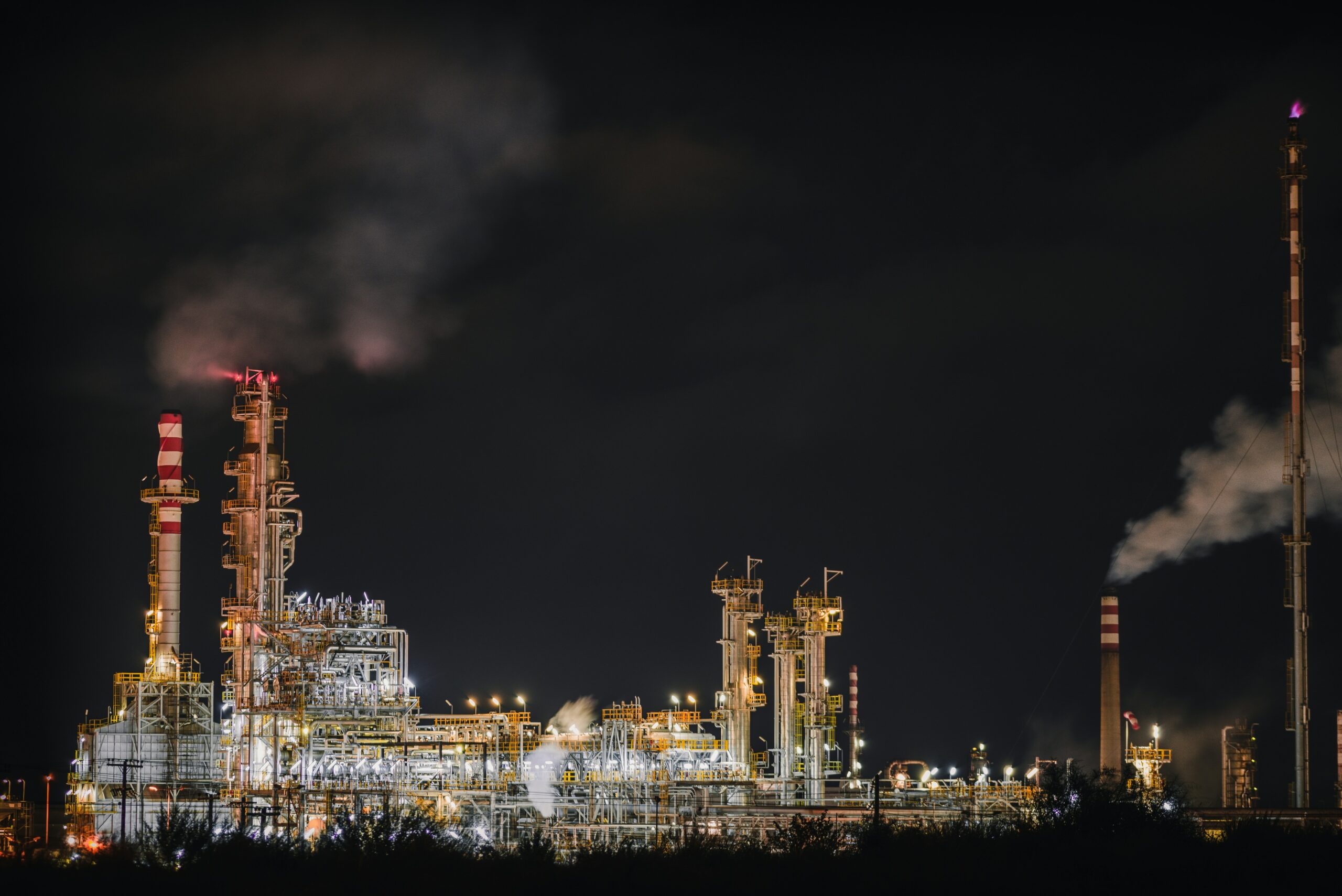 Aerial night view of a large industrial oil refinery with illuminated towers, smokestacks emitting steam, and glowing lights against a dark sky